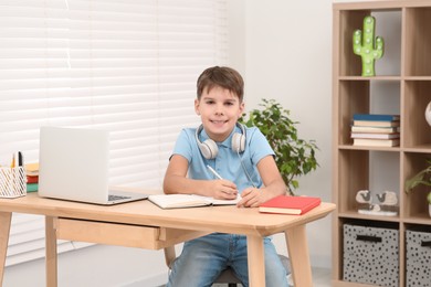 Photo of Boy writing in notepad near laptop at desk in room. Home workplace