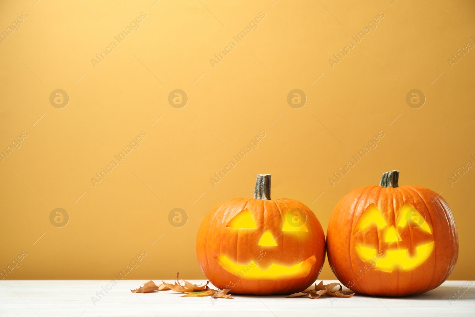 Photo of Scary jack o'lantern pumpkins and fallen leaves on white wooden table against yellow background, space for text. Halloween decor