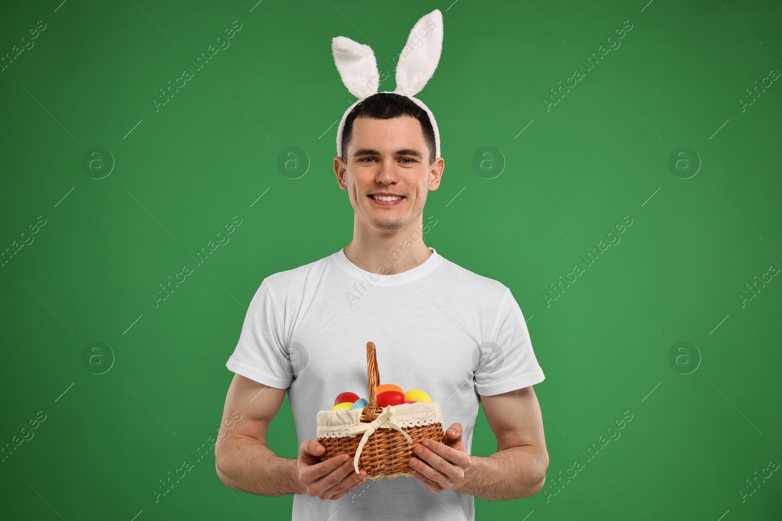 Photo of Easter celebration. Handsome young man with bunny ears holding basket of painted eggs on green background