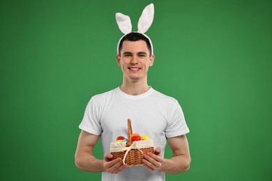 Easter celebration. Handsome young man with bunny ears holding basket of painted eggs on green background