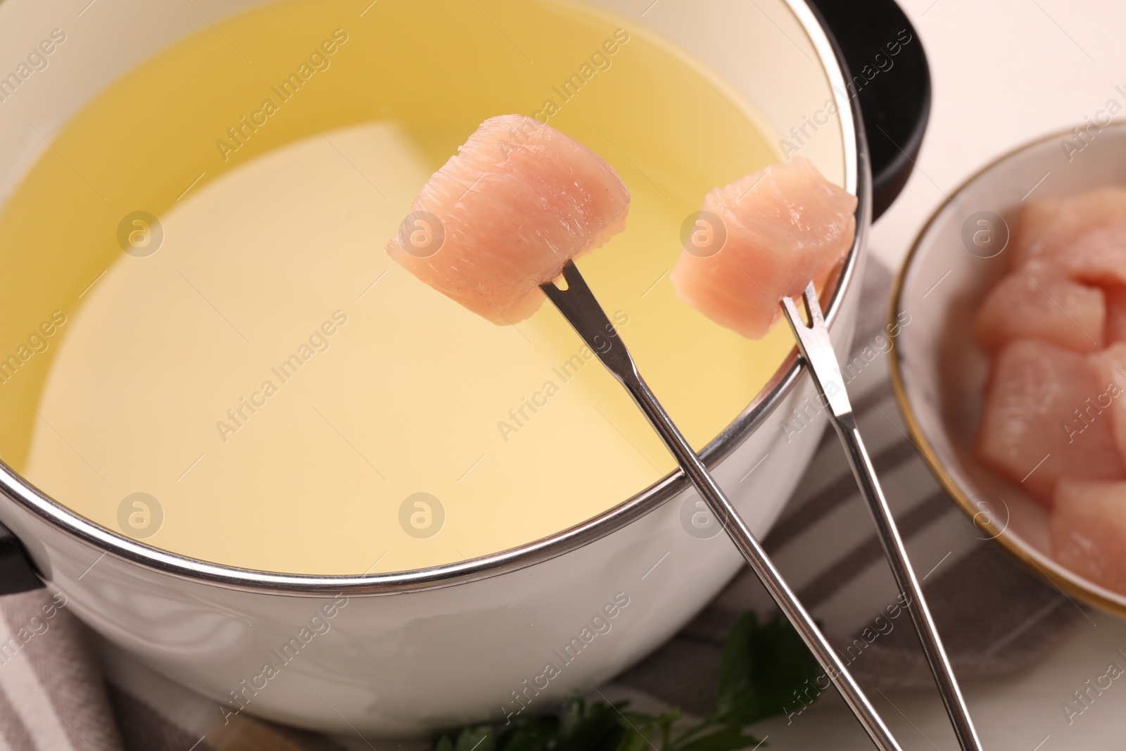 Photo of Fondue pot with oil, forks, raw meat pieces and parsley on white table, closeup