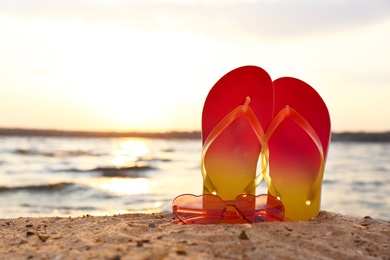 Stylish flip flops with sunglasses on sand near sea, space for text. Beach accessories