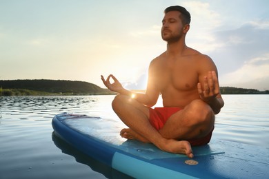 Man meditating on light blue SUP board on river at sunset