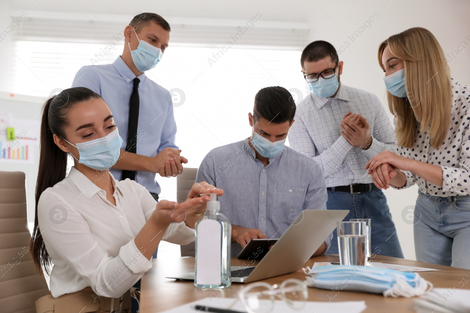 Photo of Coworkers with protective masks using hand sanitizer in office. Business meeting during COVID-19 pandemic