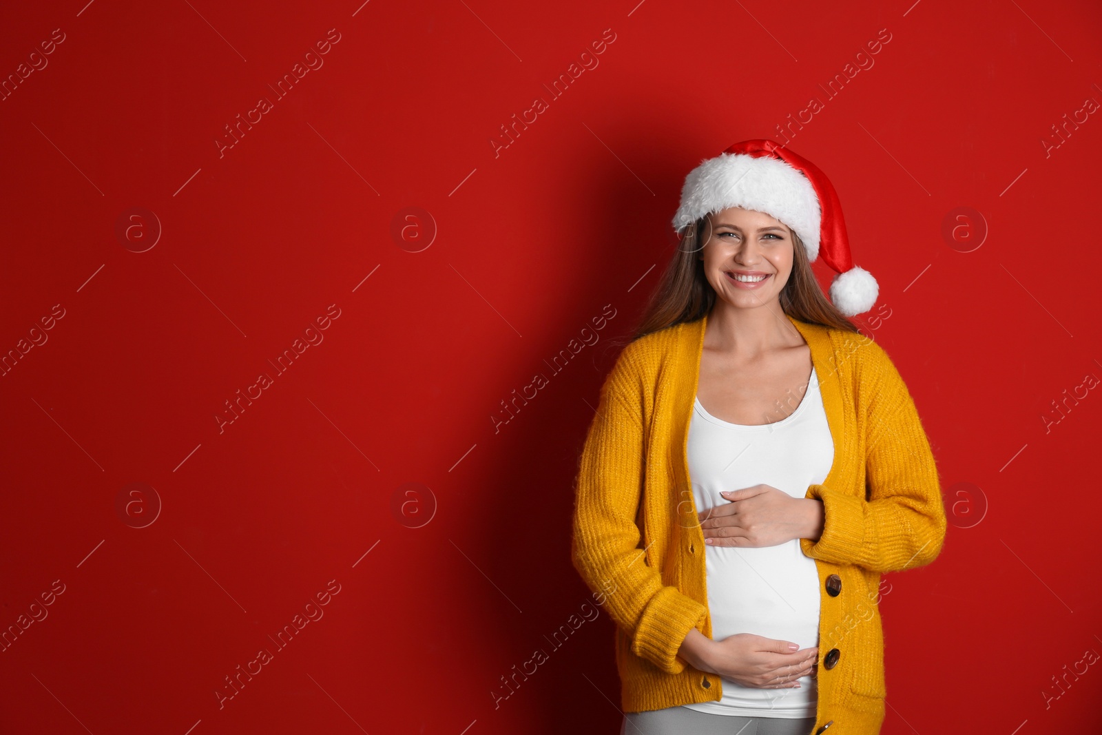 Photo of Young pregnant woman in Santa hat on red background, space for text. Christmas celebration