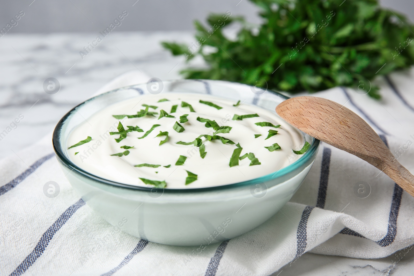 Photo of Glass bowl of fresh sour cream with parsley and wooden spoon on marble table