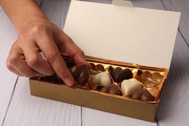 Photo of Woman taking chocolate candy out from partially empty box at white wooden table, closeup