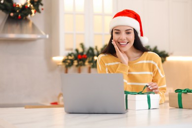 Photo of Celebrating Christmas online with exchanged by mail presents. Smiling woman in Santa hat with gift boxes during video call on laptop at home. Space for text