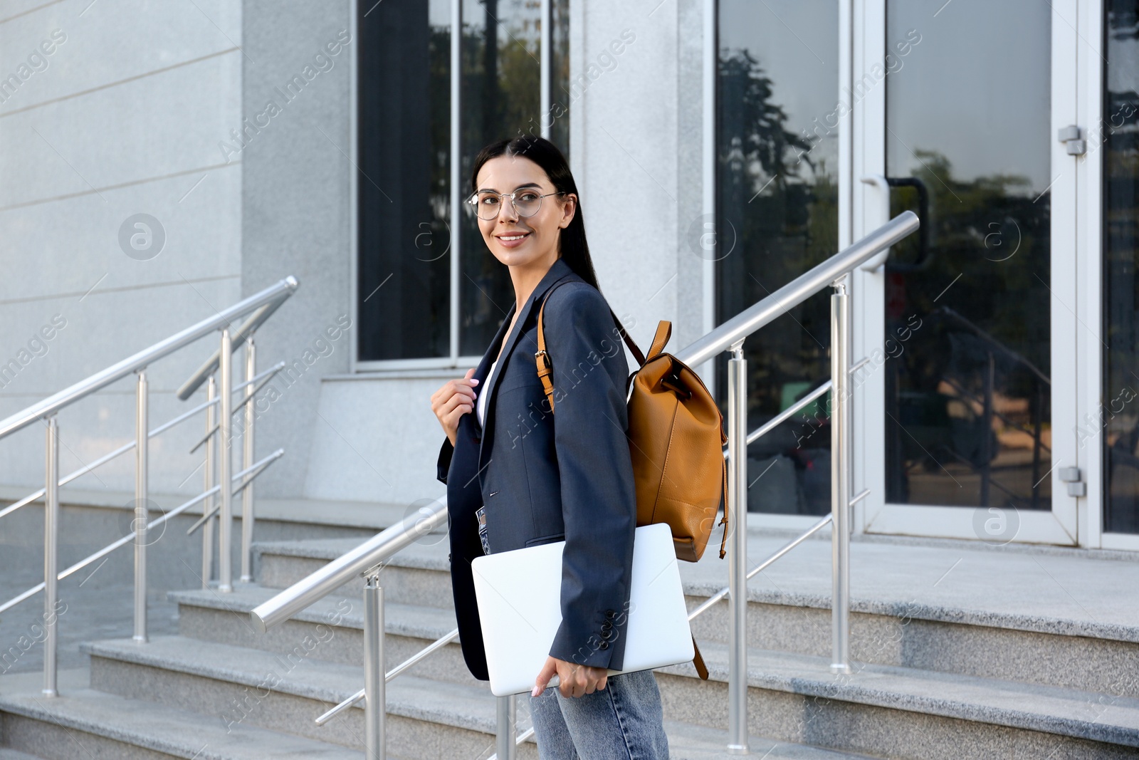 Photo of Beautiful young woman with stylish backpack and laptop near building outdoors