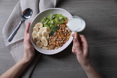 Photo of Woman pouring milk into bowl of tasty granola with banana and kiwi at grey wooden table, top view