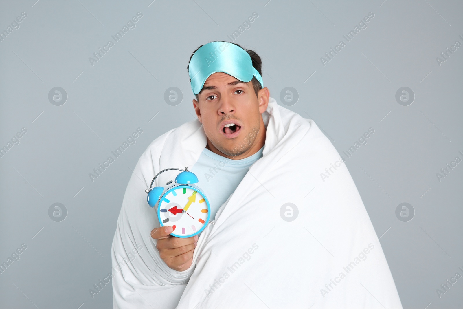 Photo of Man in sleeping mask wrapped with blanket holding alarm clock on grey background