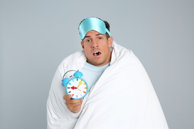 Man in sleeping mask wrapped with blanket holding alarm clock on grey background