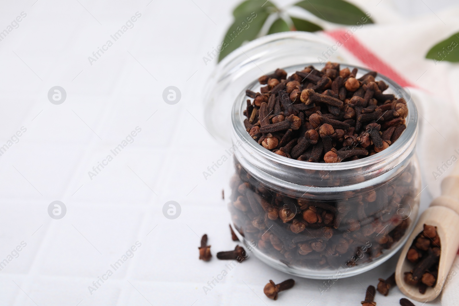 Photo of Aromatic cloves in glass jar and scoop on white tiled table, closeup. Space for text