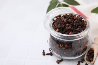 Photo of Aromatic cloves in glass jar and scoop on white tiled table, closeup. Space for text