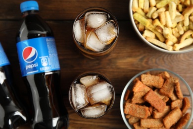 Photo of MYKOLAIV, UKRAINE - FEBRUARY 15, 2021: Glasses and bottles of Pepsi with snacks on wooden table, flat lay