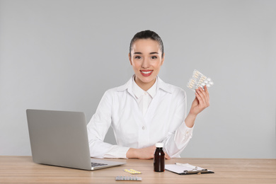 Photo of Professional pharmacist with pills and laptop at table against light grey background
