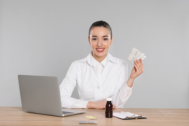 Professional pharmacist with pills and laptop at table against light grey background