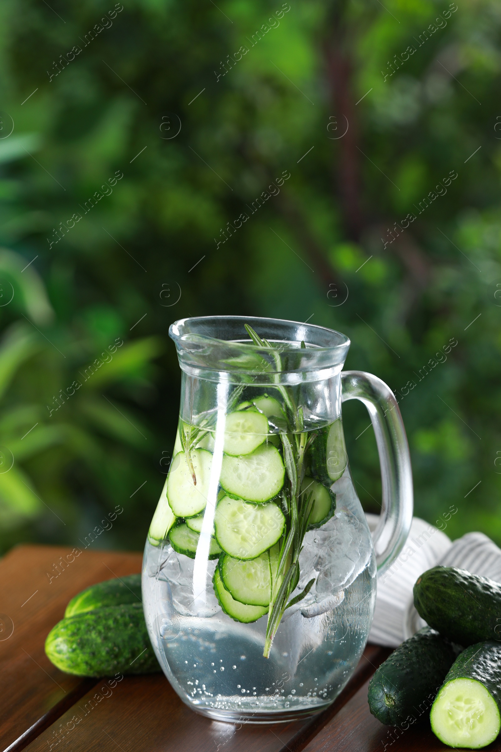 Photo of Refreshing cucumber water with rosemary in jug and vegetables on wooden table against blurred green background