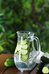 Refreshing cucumber water with rosemary in jug and vegetables on wooden table against blurred green background