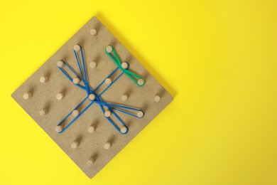 Photo of Wooden geoboard with dragonfly shape made of rubber bands on yellow background, top view and space for text. Educational toy for motor skills development