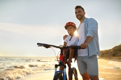 Photo of Happy father teaching son to ride bicycle on sandy beach near sea at sunset