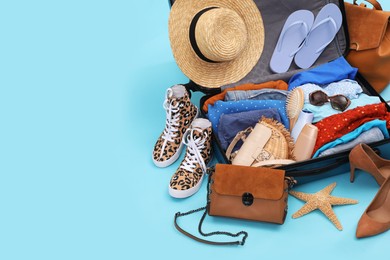Open suitcase with clothes, beach accessories and shoes on light blue background, space for text. Summer vacation