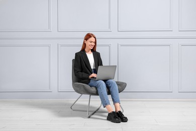 Happy young woman with laptop sitting in armchair near light grey wall indoors