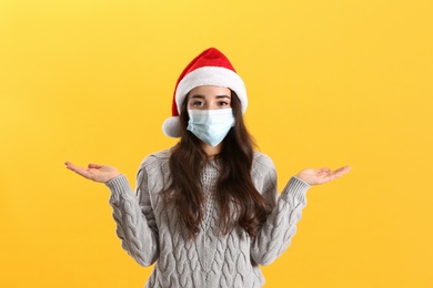 Photo of Beautiful woman wearing Santa Claus hat and medical mask on yellow background