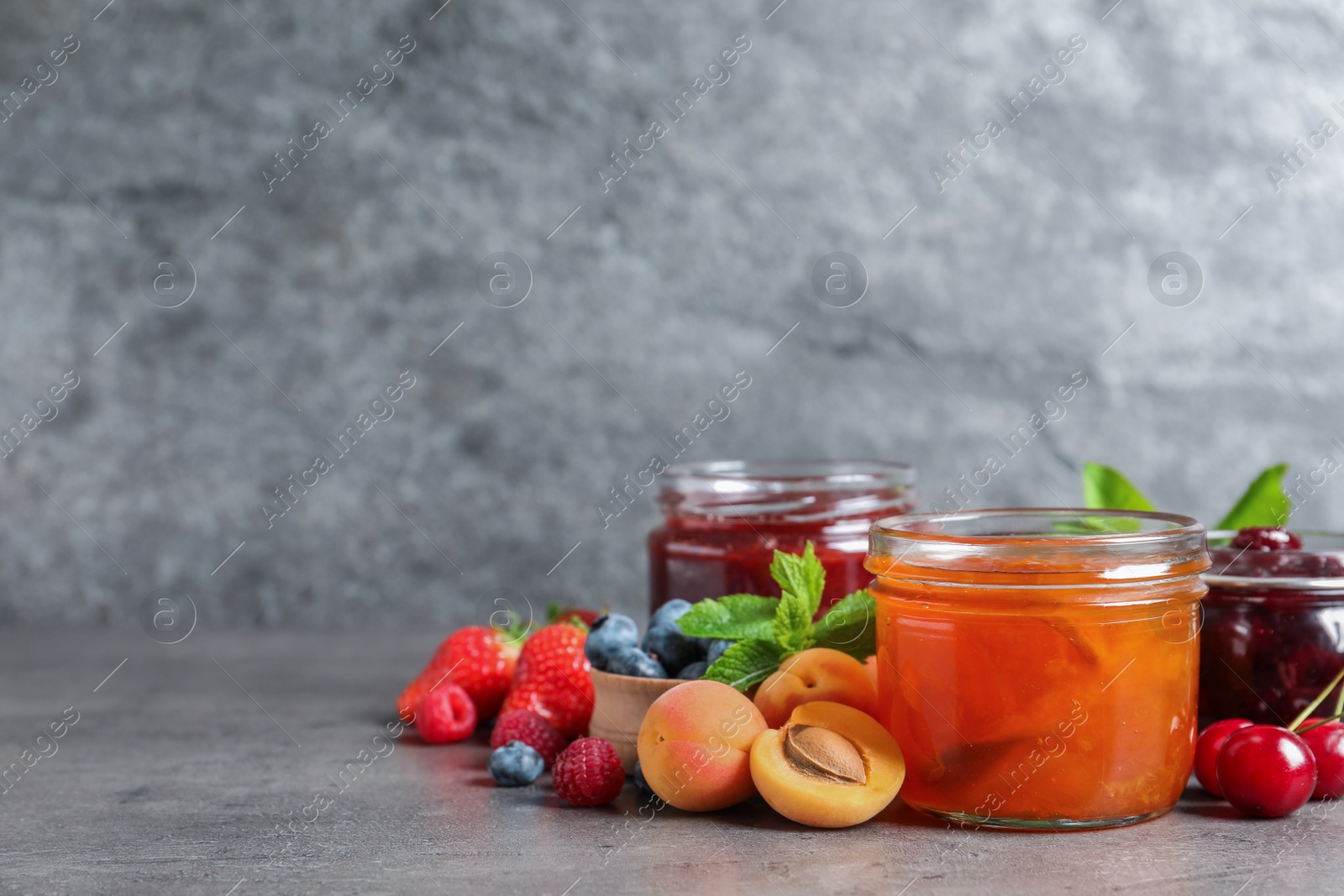 Photo of Jars with different jams and fresh fruits on grey table. Space for text