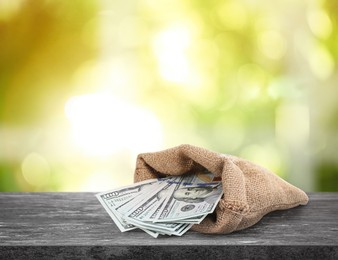 Image of Burlap bag with dollar banknotes on grey stone table against blurred green background