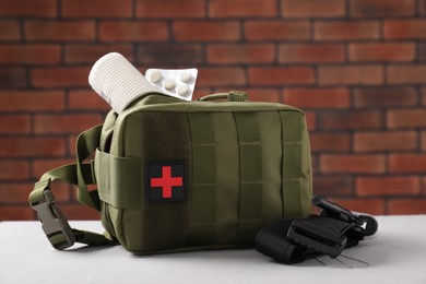 Military first aid kit, tourniquet and pills on white textured table against brick wall
