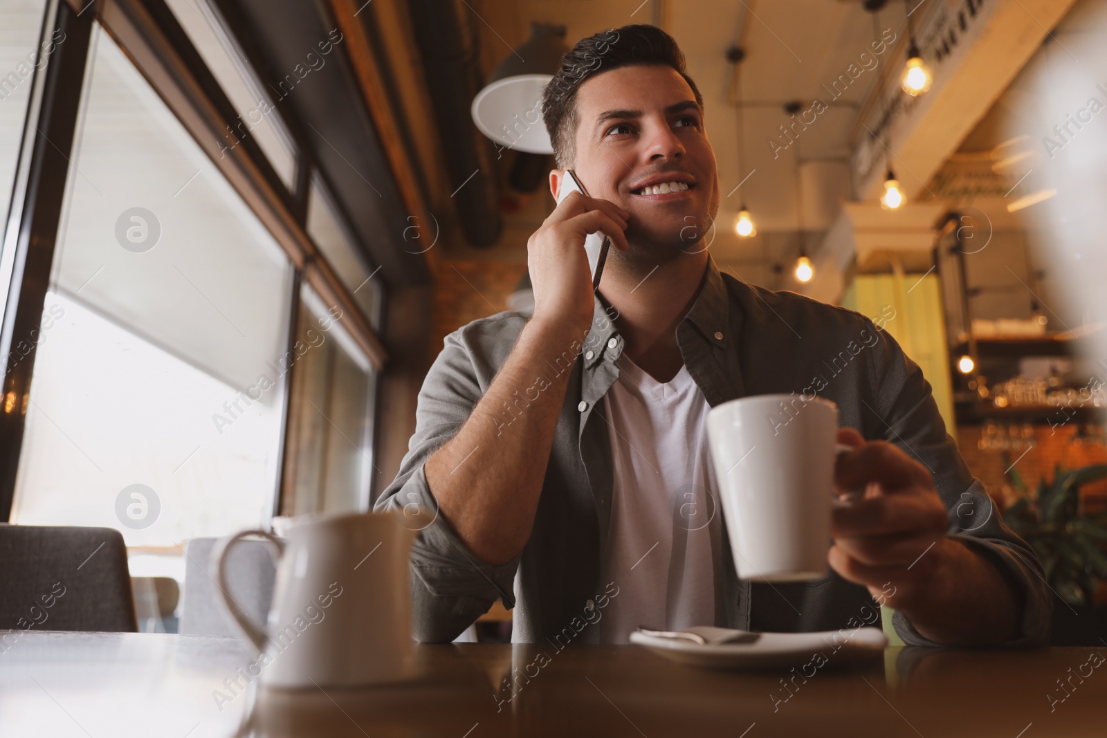 Photo of Handsome man with cup of coffee talking on smartphone at cafe in morning