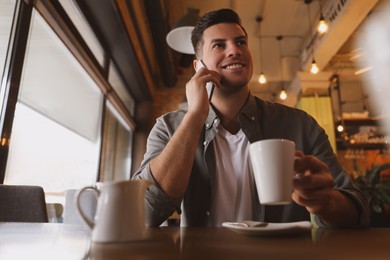 Photo of Handsome man with cup of coffee talking on smartphone at cafe in morning