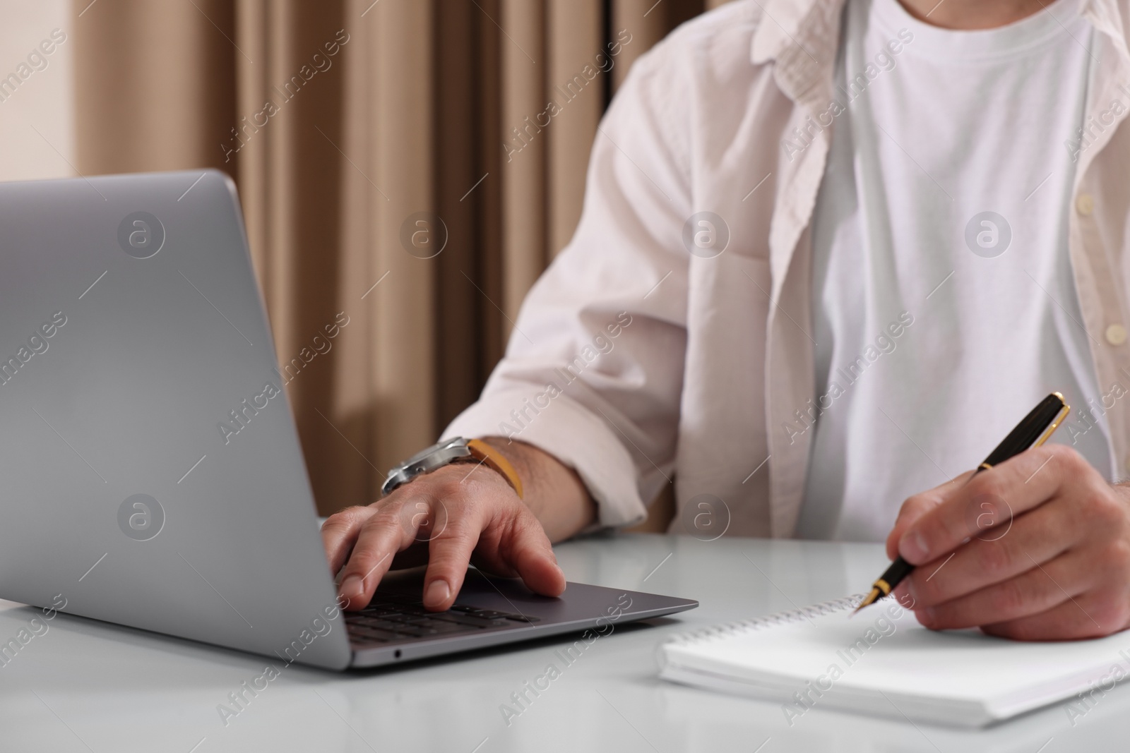 Photo of Man using laptop and writing something in notebook at table indoors, closeup