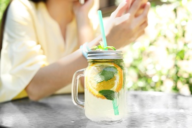 Photo of Mason jar with tasty lemonade on table and young woman at cafe, outdoors. Natural detox drink