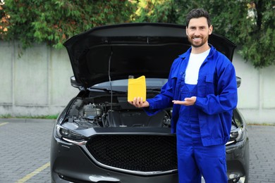 Photo of Smiling worker showing yellow container of motor oil near car outdoors