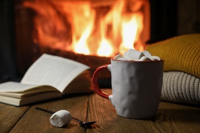Mug with hot cocoa, marshmallows and book on wooden table near fireplace