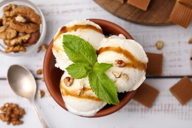 Photo of Bowl of tasty ice cream with caramel sauce, mint and nuts on white wooden table, flat lay