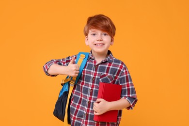 Photo of Happy schoolboy with backpack and book showing thumb up on orange background