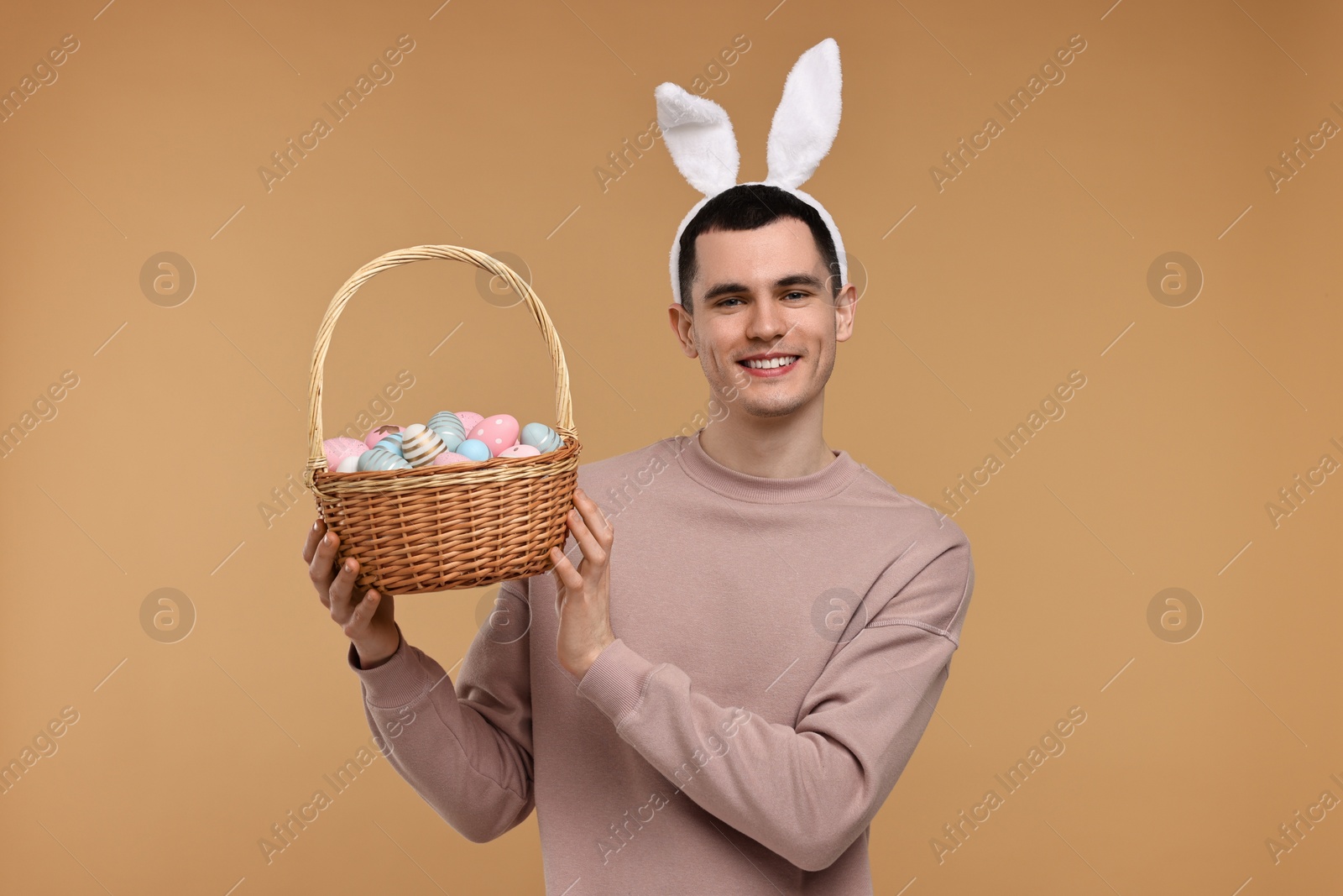Photo of Easter celebration. Handsome young man with bunny ears holding basket of painted eggs on beige background