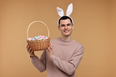 Easter celebration. Handsome young man with bunny ears holding basket of painted eggs on beige background