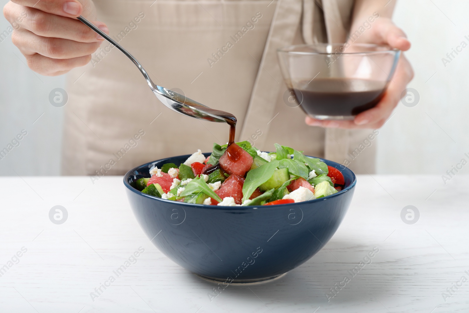 Photo of Woman adding sauce to delicious salad with watermelon, vegetables and feta cheese at white wooden table, closeup