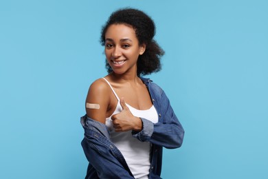 Photo of Happy young woman with adhesive bandage on her arm after vaccination showing thumb up against light blue background