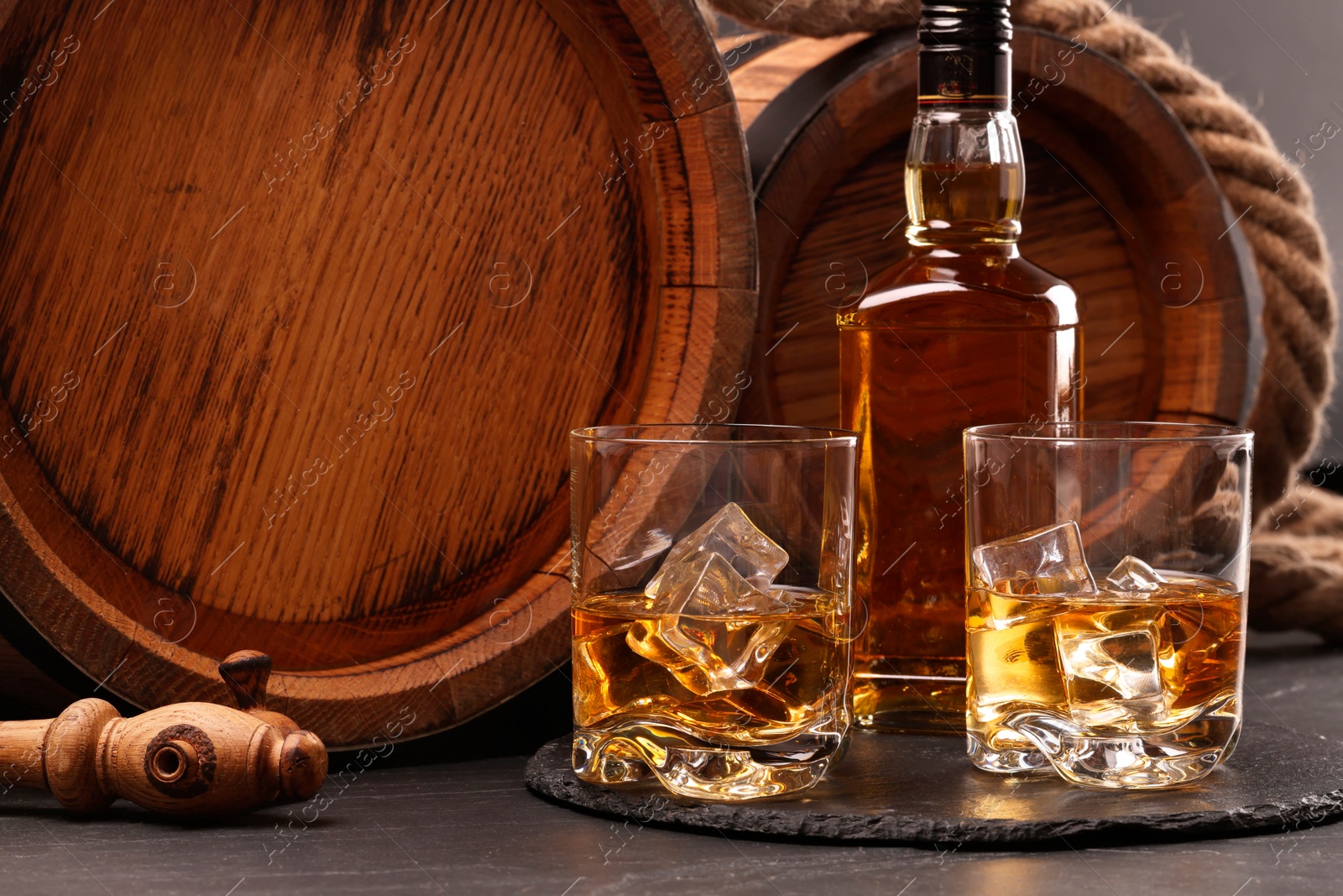 Photo of Whiskey with ice cubes in glasses, bottle and wooden barrels on black table