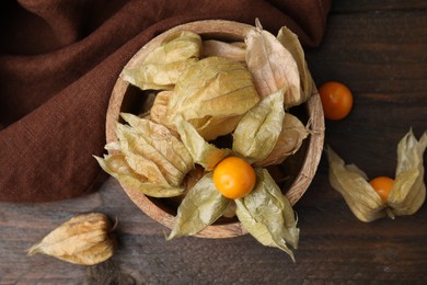 Ripe physalis fruits with calyxes in bowl on wooden table, flat lay