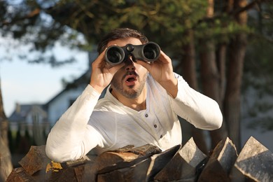 Photo of Concept of private life. Curious man with binoculars spying on neighbours over firewood outdoors