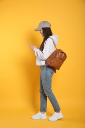 Beautiful young woman with stylish leather backpack and smartphone on yellow background