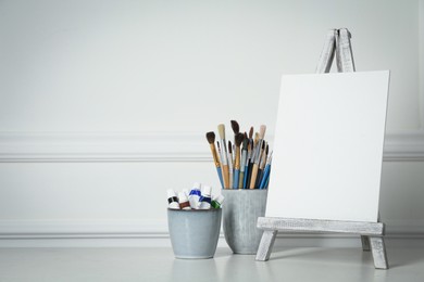 Photo of Easel with blank canvas, paints and brushes on white wooden table. Space for text