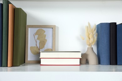 Hardcover books, picture and vase with dry flowers on shelf