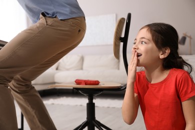 Cute little girl putting whoopee cushion on father's chair while he sitting down at home, closeup